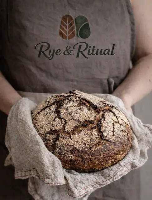 A baker in a Rye & Ritual apron holding a fresh rye loaf in a linen cloth